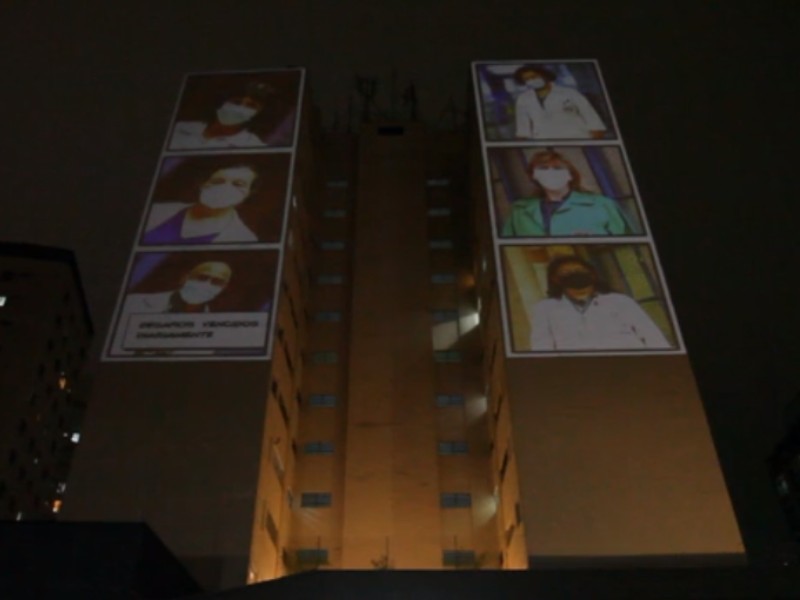 Seis fotos de profissionais da saúde são projetadas na lateral de um prédio, três na coluna da esquerda e três na da direita. As projeções foram feitas à noite e o tempo está chuvoso
